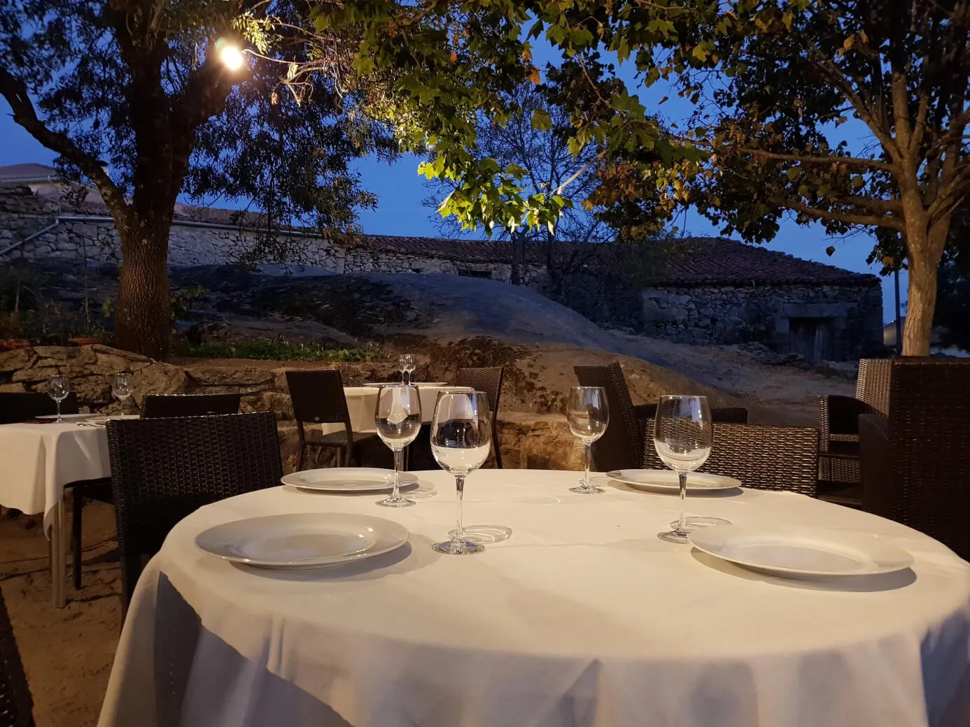 Terraza comedor de noche con mesas, sillas y árboles.